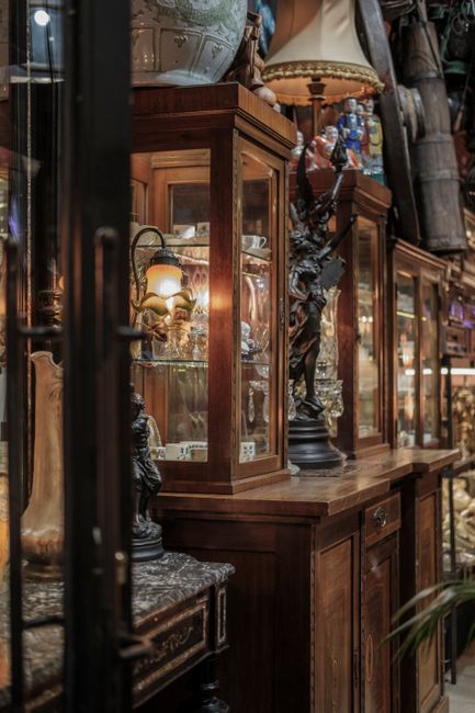 Inside a property, a large wooden shelving unit with multiple compartments displays a collection of decorative items, including a framed painting, an ornate oval mirror with a decorative gilt frame, v