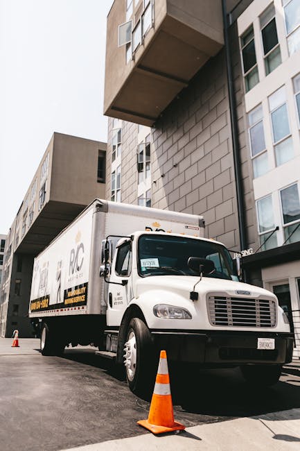 A white box truck from Man with Van Brondesbury is parked on a paved area outside a modern apartment building on Kilburn High Road. The truck is positioned in front of the building's entrance, with orange traffic cones placed nearby to manage parking. The truck's cargo area appears to be closed, ready for home relocation or furniture transport. The surrounding environment includes multi-story residential units with large windows and balconies, indicating a contemporary urban setting suitable for house removals. The overcast sky provides diffuse lighting, and the pavement is clear of additional vehicles or obstructions. The scene captures the loading process typical of professional removal services, such as those offered by [COMPANY_NAME], during packing and moving operations in an inner-city area.
