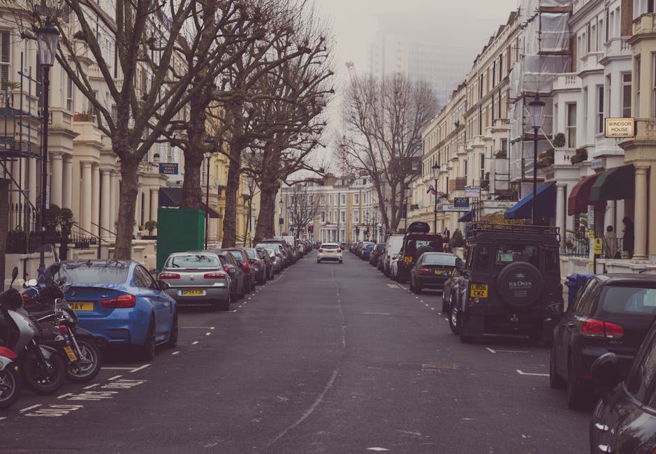 A wide residential street with rows of multi-storey Victorian-style terraced houses on both sides, featuring decorative bay windows, stoops, and small front gardens. The street is lined with leafless trees, indicating a winter or early spring setting, with parked cars of various makes and models including sedans, hatchbacks, and SUVs along both curbs. On the right side, a black off-road vehicle and a row of compact cars are visible, while on the left, a blue vehicle and several other cars are parked. A few motorbikes are also parked towards the front left corner. The pavement is clean, and the road appears to be in good condition with clear parking spaces marked. Some of the houses have small awnings or canopies over their entrances, and traditional street lamps are mounted on the house facades. The scene captures a typical urban residential area suitable for home relocation or furniture transport, with no moving activity currently visible but implying a setting where professional removals like those by Man with Van Brondesbury could operate to avoid parking fines during house moves on Kilburn High Road, BRONDESBURY.