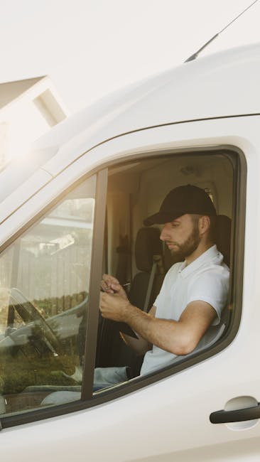 A man with a beard wearing a black cap, white t-shirt, and jeans is lifting a cardboard box from the back of a white van during a home relocation process. Inside the van, there are several other cardb