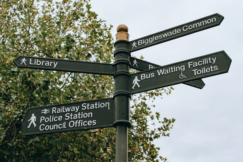 A black multi-directional street signpost with six directional signs pointing towards various locations including Biggleswade Common, Library, Bus Waiting Facility and Toilets, Railway Station, Police Station, and Council Offices, mounted on a metal pole with a rounded top. The signs have white text and icons, with some including walking symbols and wheelchair accessibility icons. The background features a leafy tree with green and yellow foliage under a cloudy sky, indicating an outdoor setting. The scene is relevant to local navigation, transportation, and public facilities, supporting house removals and moving services by providing contextual outdoor environment details. Images like this are useful for understanding local infrastructure and logistics for home relocations, as seen on the webpage for Brent Council Permits for Removals in Brondesbury by Man with Van Brondesbury.