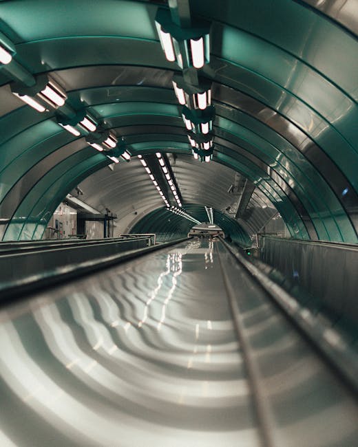 A wide view inside an underground escalator tunnel with a curved ceiling featuring multiple rows of bright, evenly spaced LED lights. The escalator handrails are visible in the foreground, extending into the distance, leading towards the tunnel’s exit or continuation. The tunnel’s exterior walls are clad in smooth, reflective greenish-blue panels, creating a modern, clean appearance. The scene is illuminated with artificial lighting, highlighting the tunnel structure and giving a sense of movement and depth. This setting relates to transportation or transit environments that can be part of house removals or home relocation logistics, as handled by Man with Van Brondesbury, ensuring safe, professional furniture transport and packing during moving processes.