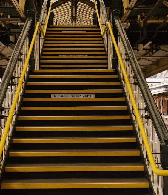 Inside a busy underground station with curved, tunnel-like walls and a ceiling fitted with long, fluorescent light fixtures. Multiple people are standing on an upward-moving escalator, some looking at their phones while others face forward. The escalator features black handrails and metal steps, with a yellow safety line at the edge. To the sides, there are a series of framed posters or maps attached to the wall panels. In the background, additional passengers can be seen walking at the top and bottom of the escalator, with stairs visible beyond the top. The scene is illuminated with neutral, slightly dim lighting typical of underground stations, depicting a moment during a home relocation or moving process where people are commuting, and transport logistics are involved, relevant to furniture transport and packing and moving logistics at Brondesbury Station, as managed by Man with Van Brondesbury.