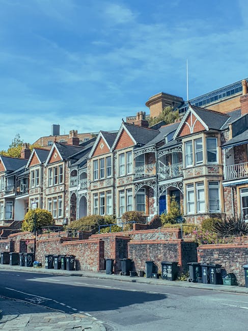 Photograph of a row of Victorian-style terraced houses in Brondesbury, featuring ornate ironwork balconies and bay windows. The buildings are constructed with brick and painted wood detailing, with sloped tiled roofs. The houses are elevated above the street level, accessed by steps, with small landscaped gardens and shrubbery in front. A clear blue sky and daylight illuminate the scene. At street level, there is a pavement with several black wheelie bins aligned along the curb. This residential setting exemplifies typical house architecture suitable for home relocation and moving services, and it provides context for local property types handled by companies like Man with Van Brondesbury during furniture transport and packing and moving operations.
