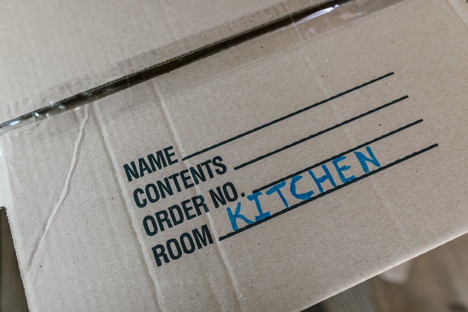 Close-up view of a cardboard moving box with a printed label on the side, showing fields for name, contents, order number, and room. The room field is handwritten in blue ink with the word 'KITCHEN'. The box is situated inside a home, likely near an entry or in a storage area, indicating the packing process during a home relocation or furniture transport. The cardboard is sealed with transparent packing tape, and the label is neatly printed, emphasizing organized packing and moving logistics. The environment is well-lit, and other boxes or packing materials may be partially visible in the background, reflecting the typical scene during professional removals carried out by companies such as Man with Van Brondesbury.