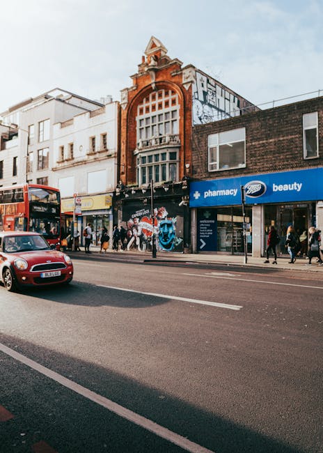 A street scene on Kilburn High Road showing a row of commercial buildings including a pharmacy and a beauty shop, with pedestrians walking along the pavement. A red double-decker bus and a small red car are visible on the road, along with a white van parked near the curb. In the background, an ornate brick building with a large arched window and decorative architectural details stands above the other shops. The scene captures a typical urban environment suitable for home relocation or furniture transport services, with visible road markings, storefront signage, and people engaged in daily activities. Man with Van Brondesbury’s removal service could assist with loading household items into nearby vehicles, facilitating efficient house removals and packing processes in this busy area.