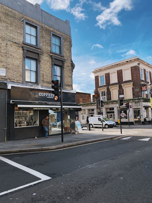The image depicts a street scene on Kilburn High Road showing a two-storey building with a black storefront labeled 'COFFEE' on the ground floor, featuring large windows and a person inside near the counter. In front of the shop, a pedestrian stands on the sidewalk, waiting or observing. Opposite the shop, there is a white van parked in a loading bay area with a red traffic light visible on a pole next to it. The van appears to be involved in a home relocation or furniture transport process, likely related to a house removals service such as Man with Van Brondesbury, as part of a typical moving logistics operation. The street has marked parking spaces and a clear blue sky with some scattered clouds, indicating daytime. Nearby buildings include a traditional brick building to the right with decorative architectural details and large windows, emphasizing an urban environment suited for moving activities.