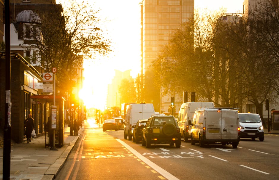 A busy urban street scene during sunset featuring a line of parked and moving vehicles including white vans and cars, positioned along the right side of the road with some vehicles loading or unloading. In the foreground, a closed bus shelter with a red London Underground sign and timetable poster is situated on the pavement. Pedestrians are visible walking along the sidewalk, some near the shelter. Tall buildings and trees line both sides of the street, with one building on the left having a rounded dome on its roof. The sunlight filters through the trees and reflects off the vehicles, creating a warm, golden glow across the scene which suggests ongoing home relocation or furniture transport activities undertaken by a professional moving service, such as Man with Van Brondesbury, in an area like Willesden Lane (NW6). The environment appears calm with a clear sky and well-maintained surroundings, capturing the typical logistical aspects of house removals involving loading and packing in an urban setting during late afternoon or early evening.