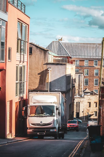 A white box truck parked on a narrow urban street next to multi-storey residential buildings with red and gray exteriors, some with balcony windows. The truck is positioned partly on the pavement and partly on the road, with its cab facing the camera. The scene shows the loading process of household furniture and boxes for home relocation, with the street that appears to be in a residential neighbourhood in Brondesbury, NW6. Nearby, two cars, including a black vehicle, are parked along the curb. The sky is partly cloudy with natural daylight illuminating the scene, highlighting the details of the buildings, the truck, and adjacent street environment, which is consistent with moving logistics managed by Man with Van Brondesbury, specialising in removals and furniture transport.