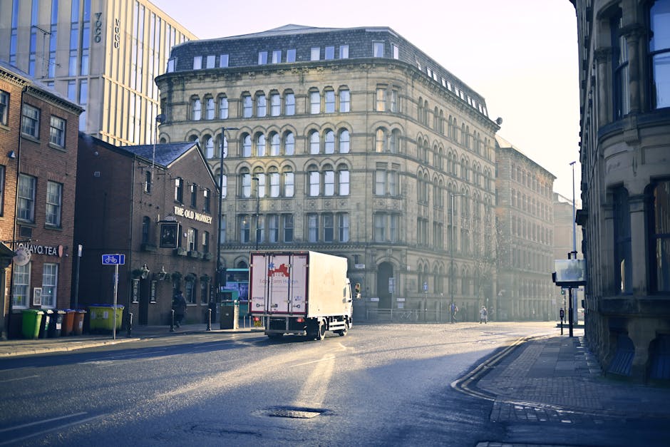 A white moving van is parked on a city street in Brondesbury during daytime, with its rear doors open for loading or unloading. Inside the van, several cardboard boxes, wrapped furniture pieces, and packing materials such as bubble wrap and blankets are visible, prepared for a home relocation. A person is seen carrying a large, wrapped item toward the van, with other packing supplies nearby. The surrounding environment includes a historic multi-storey sandstone building with multiple arched windows, modern adjacent buildings, and a few pedestrians walking on the pavement. The street is relatively quiet with minimal traffic, and additional moving equipment like trolleys and straps are nearby, indicating a professional furniture transport or packing and moving process. Man with Van Brondesbury specializes in efficient removals, and this scene captures part of their loading process at a central NW6 location.