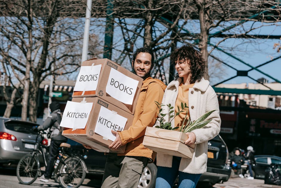 A man and a woman are engaged in a home relocation task outdoors during daylight, carrying packed cardboard boxes labeled 'BOOKS' and 'KITCHEN' in large, clear font, indicating they contain household items being prepared for moving. The man holds two cardboard boxes stacked in his arms, which are secured with packing tape and have printed labels, signifying careful packing and organising for furniture transport. The woman carries a wooden crate filled with potted plants, showcasing the wrapping materials used for fragile items. Both are dressed casually in jackets suitable for cool weather, and they are smiling as they progress along a paved area with parked cars and bicycles in the background, suggesting an active busy moving day. The background features leafless trees and a partially visible modern building structure, indicating an urban environment. This scene illustrates the practicality of loading and packing processes during a house removal, supported by the services of [COMPANY_NAME], such as packing and moving logistics, displaying the importance of careful handling and transportation of household belongings as part of a professional removals operation.
